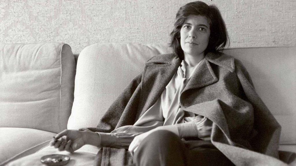 Susan Sontag recostada en una cama, vistiendo un saco claro sobre blusa blanca, mirando hacia un lado
