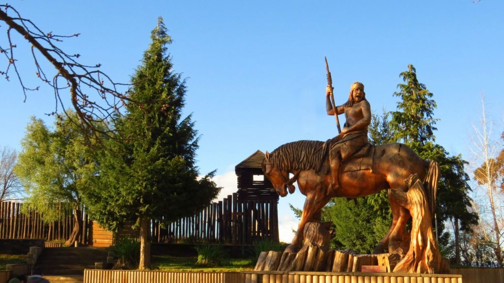 Estatua de bronce de Pelantaro a caballo con lanza levantada, rodeada de árboles y cerca de madera