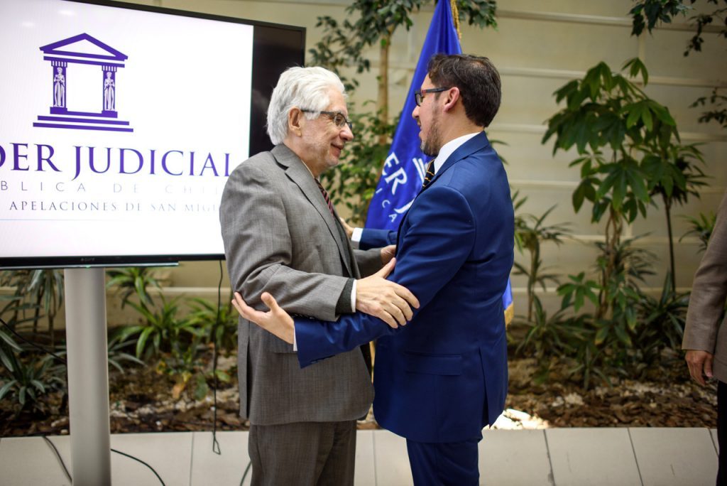 Dos hombres en traje saludan cordialmente frente a banner del Poder Judicial, en acto académico