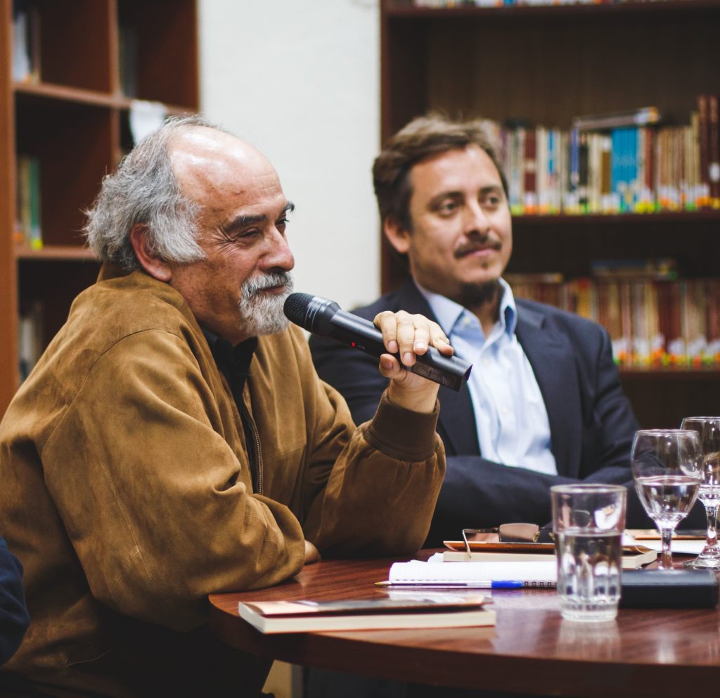 Dos hombres en mesa de panel: uno mayor con gafas habla al micrófono; otro más joven escucha. Biblioteca de fondo