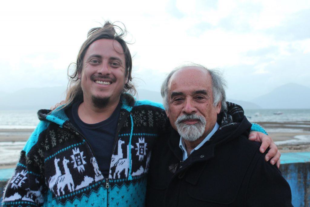 Dos hombres posando junto al mar, uno con cabello largo y chaqueta azul, otro mayor con barba gris y chaqueta negra