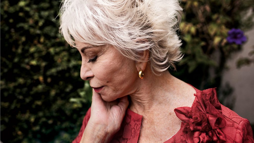 Mujer de cabello blanco ondulado, vistiendo blusa roja con flor decorativa, en perfil mirando hacia abajo