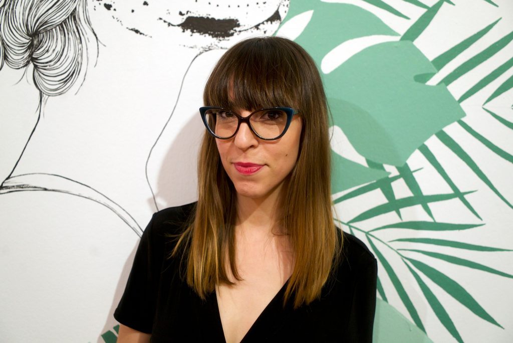 Retrato de Laura Agustí, ilustradora y autora, con gafas y cabello oscuro frente a ilustraciones de trazo verde