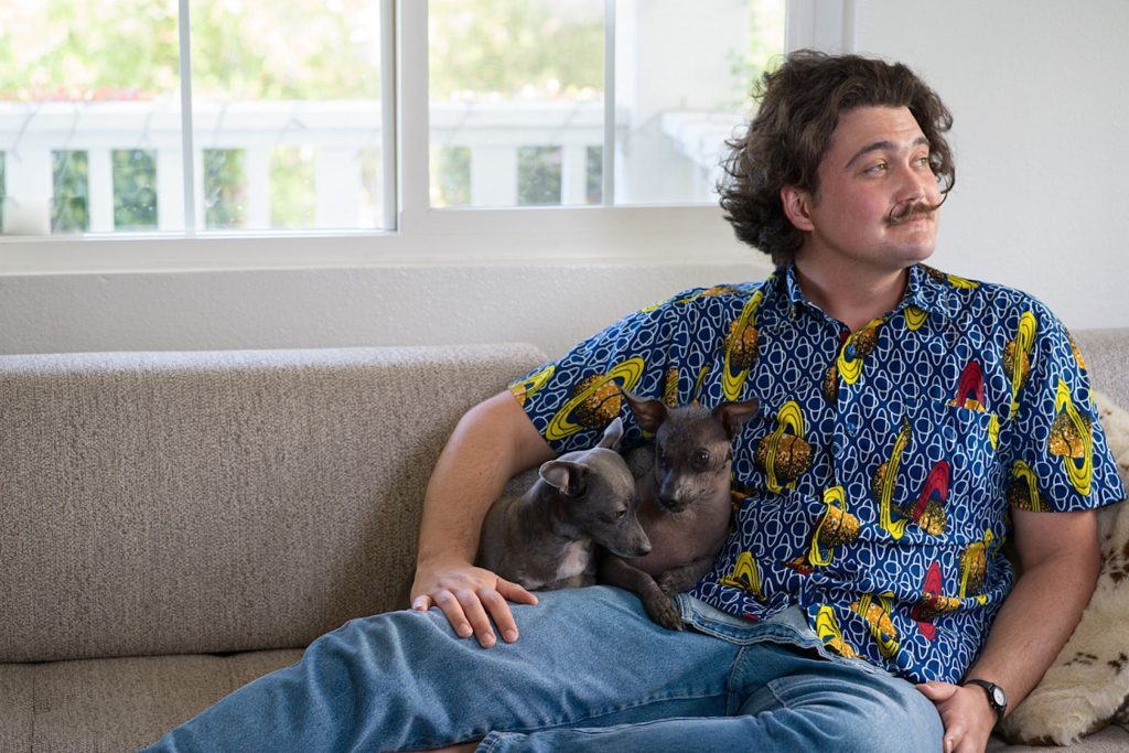 Hombre sentado en sofá gris sosteniendo a un gato gris, vistiendo camisa colorida con estampado geométrico azul