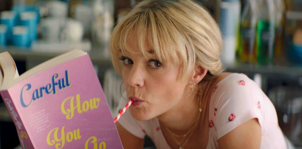 Mujer rubia con libreta rosa titulada Careful How You Go en una habitación con estanterías de fondo