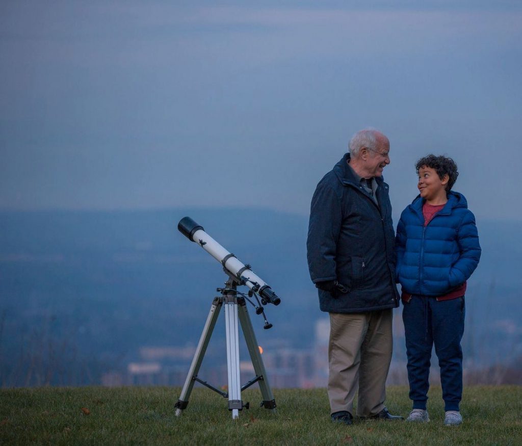 Dos personas observan el cielo nocturno a través de un telescopio en una colina con vista panorámica al atardecer