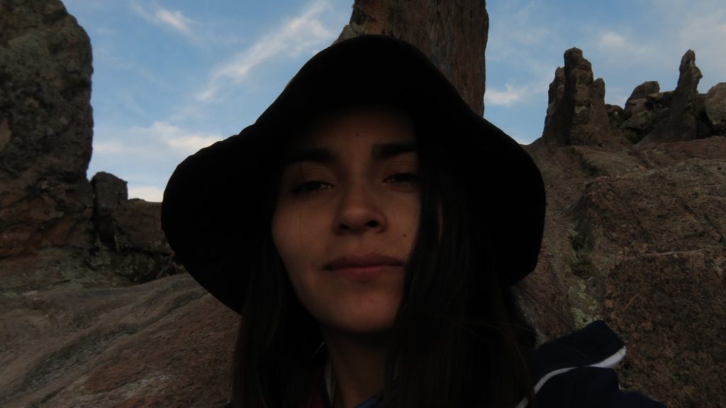 Mujer de cabello largo y oscuro con capucha negra, ruinas de piedra de fondo y cielo nublado