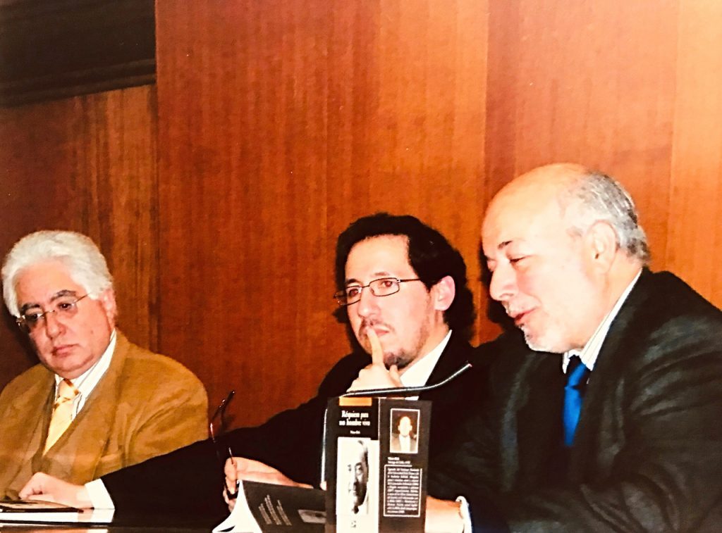 Tres hombres en traje durante panel o conferencia, el central con micrófono, fondo de madera naranja