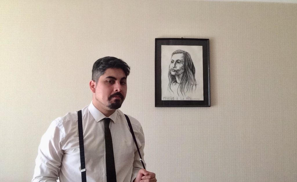 Nicolás López-Pérez, escritor, de pie frente a retrato enmarcado, vistiendo camisa blanca y corbata oscura