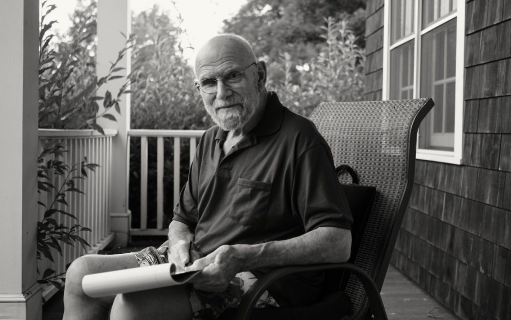 Oliver Sacks sentado en una terraza, con gafas y camisa oscura, sosteniendo un libro en sus manos