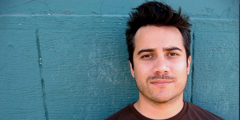 Hombre joven con cabello negro despeinado frente a pared azul turquesa, sonriendo levemente hacia la cámara