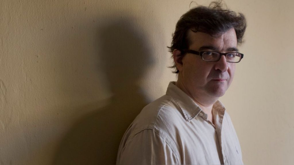 Javier Cercas, escritor español, de perfil contra pared blanca con gafas y camisa clara