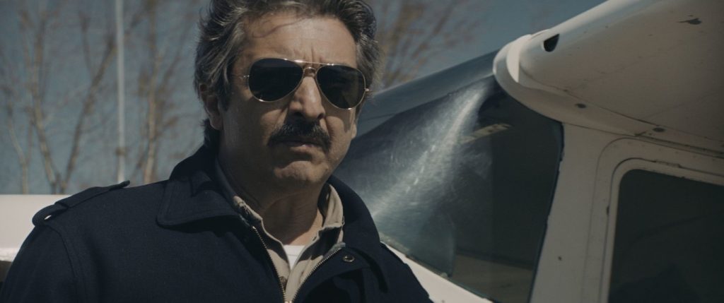 Hombre de mediana edad con gafas de sol y bigote junto a una camioneta blanca en escena de tensión