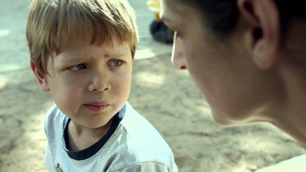 Niño rubio con camiseta blanca mira a un adulto de perfil en escena de drama familiar al aire libre