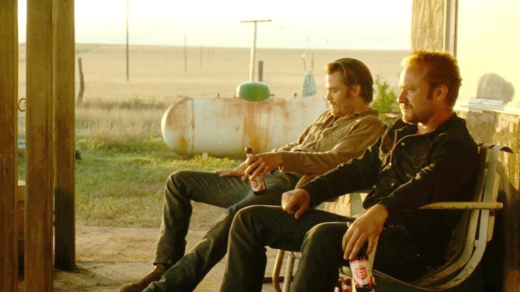Dos hombres sentados en sillas en un porche, observando el paisaje rural de las llanuras al atardecer