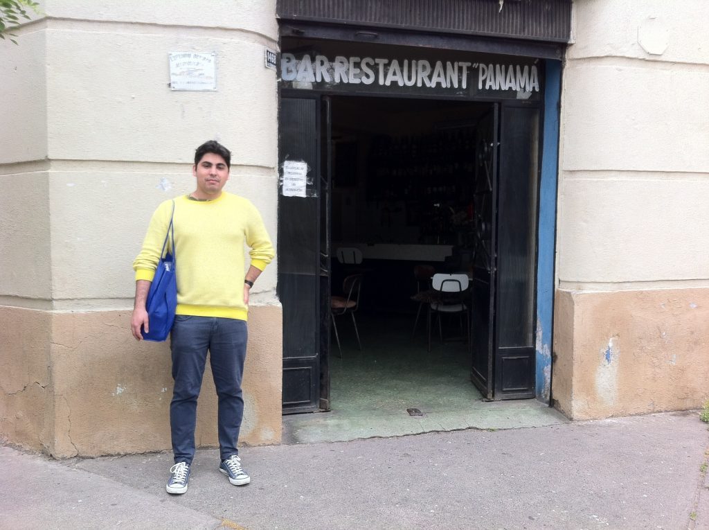 Nicolás López Pérez de pie frente a la entrada de un bar restaurante, vistiendo suéter amarillo y chaqueta azul