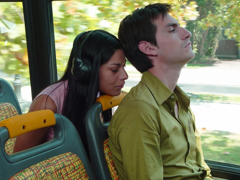 Dos personas en un autobús: mujer con auriculares y hombre de camisa verde mirando por la ventana hacia un parque