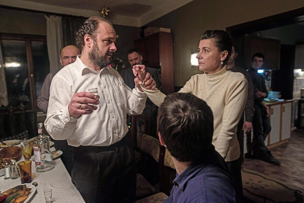 Hombre de camisa blanca gesticulando durante conversación animada en interior, con otras personas alrededor en ambiente