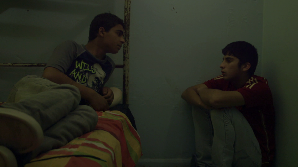 Dos jóvenes sentados en el piso de una habitación con paredes verdes, conversando en ambiente de penumbra