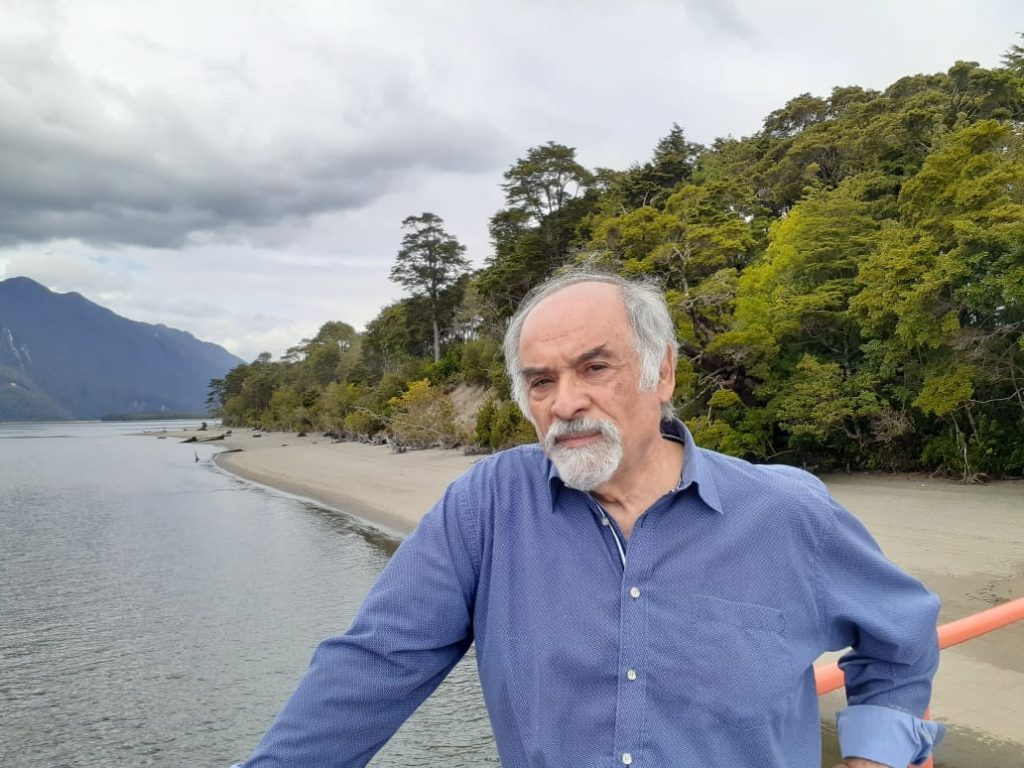 Juan Mihovilovich frente a lago con bosque y montañas al fondo, vistiendo camisa azul