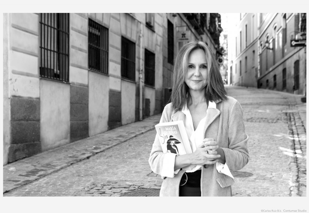 Mujer con blazer blanco en calle empedrada de ciudad antigua, mirando a cámara con expresión seria y segura