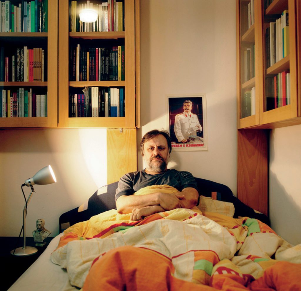 Slavoj Žižek sentado en una cama rodeado de libros y revistas, con expresión pensativa en su habitación