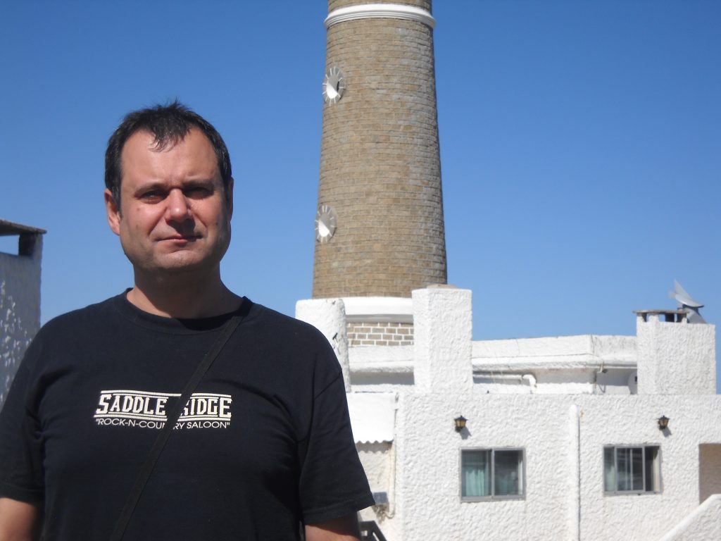 Aníbal Ricci, escritor chileno, frente a un faro blanco bajo cielo despejado, vistiendo camiseta negra