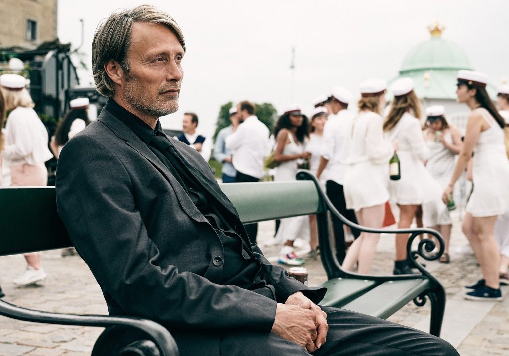 Mads Mikkelsen sentado en un banco de madera, mirando hacia la cámara, con personas al fondo en un espacio público