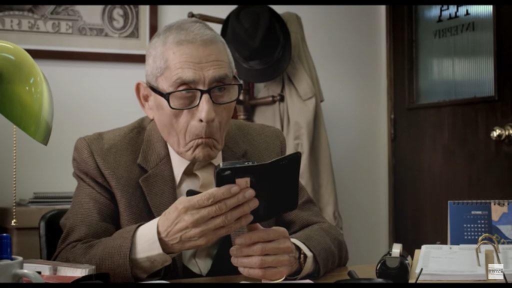 Hombre mayor con gafas y traje marrón sostiene una tablet en un escritorio con documentos, en escena de intriga