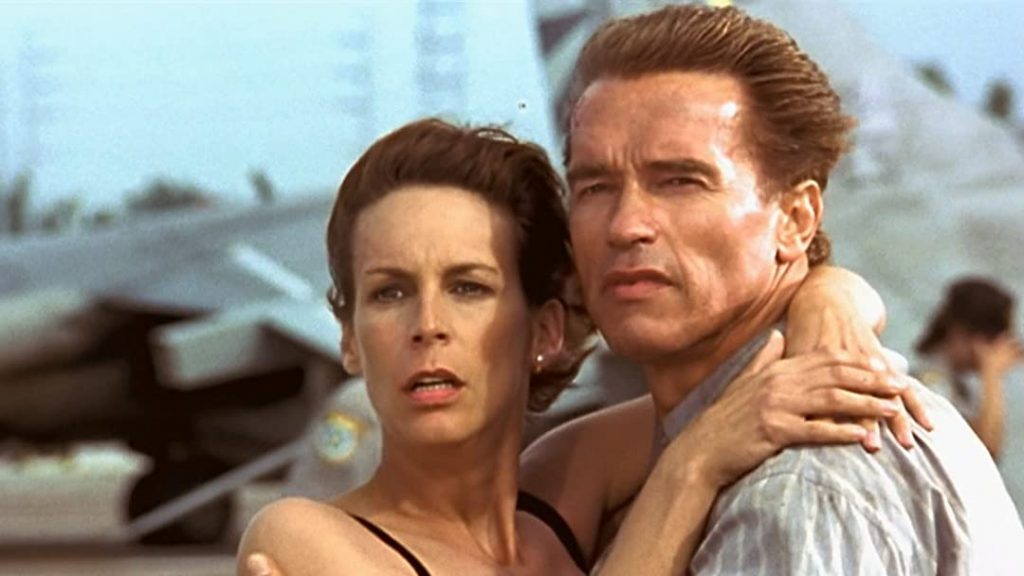 Pareja en escena de acción con avión al fondo, ella en vestido claro y él abraza protectoramente durante momento