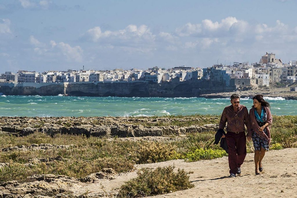 Dos personas caminan por una playa desértica hacia una ciudad costera con edificios al fondo y mar turquesa