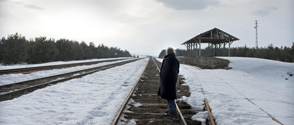 Hombre solitario caminando sobre vías de tren nevadas hacia una estación de madera en paisaje invernal desolado
