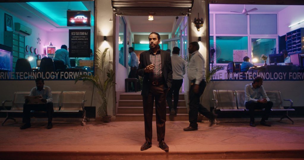 Actor protagonista en escena de tensión urbana, con personajes en fondo desenfocado y iluminación neon azul y púrpura