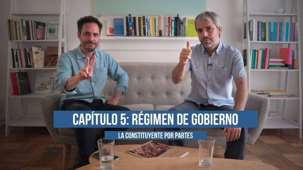 Dos hombres en estudio conversan frente a cámara junto a escritorio, con libreros de fondo y banner azul con texto