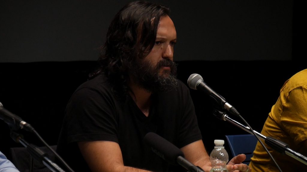 Hombre con cabello largo y oscuro en panel de discusión, micrófono frente a él, fondo oscuro