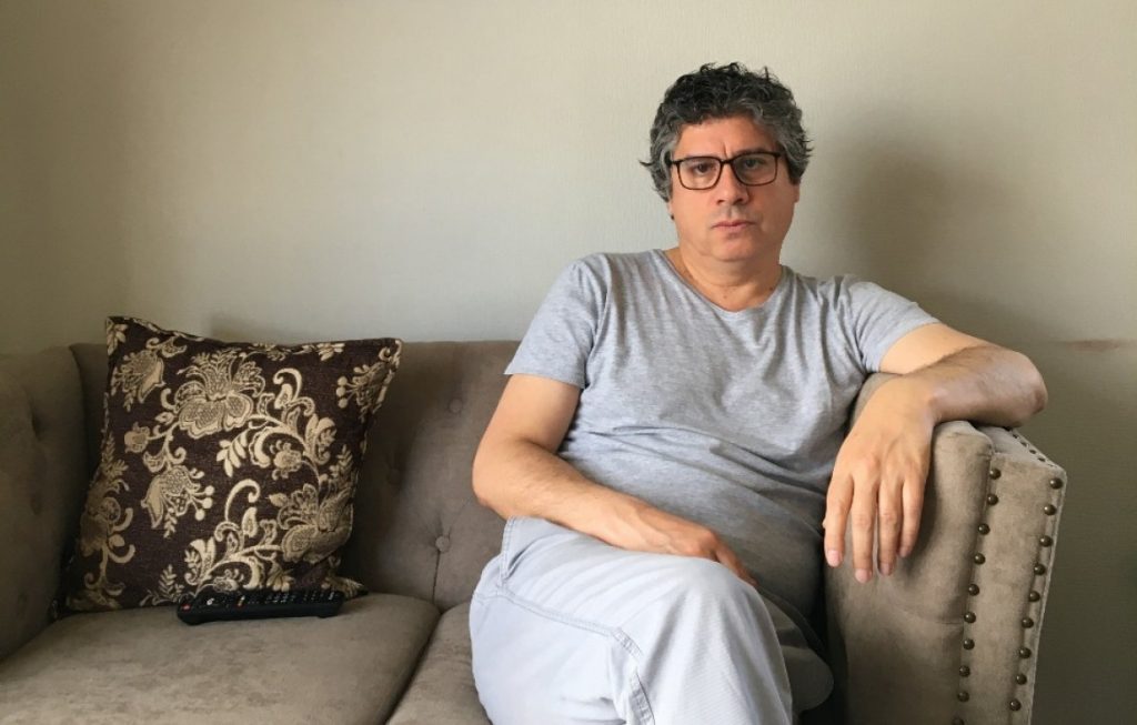 Ricardo Herrera Alarcón, poeta, sentado en sofá gris con cojines estampados, vistiendo camiseta azul claro