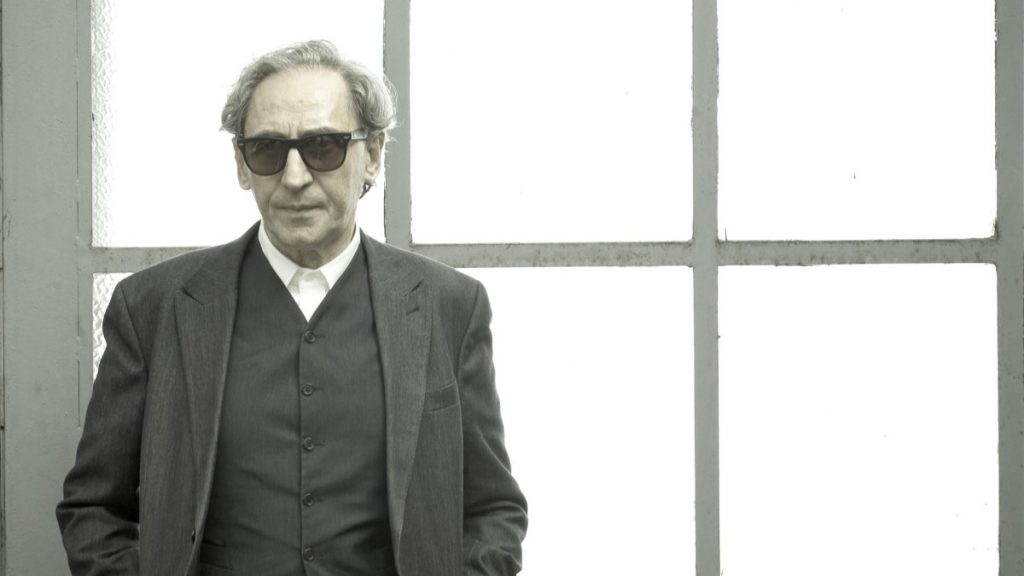 Franco Battiato con gafas de sol y traje oscuro frente a una ventana con marcos blancos