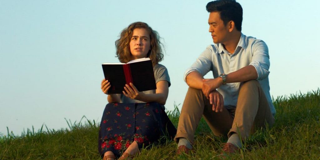 Pareja joven sentada en el pasto leyendo un libro juntos bajo cielo despejado en escena de la película Columbus