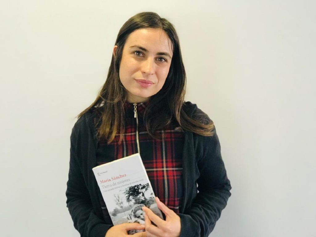 Mujer sostiene libro Tierra de mujeres, viste cardigan negro y bufanda escocesa roja, fondo blanco neutro