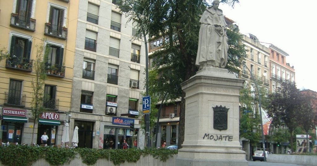 Estatua de Tirso de Molina en plaza urbana rodeada de edificios antiguos y vegetación en Madrid