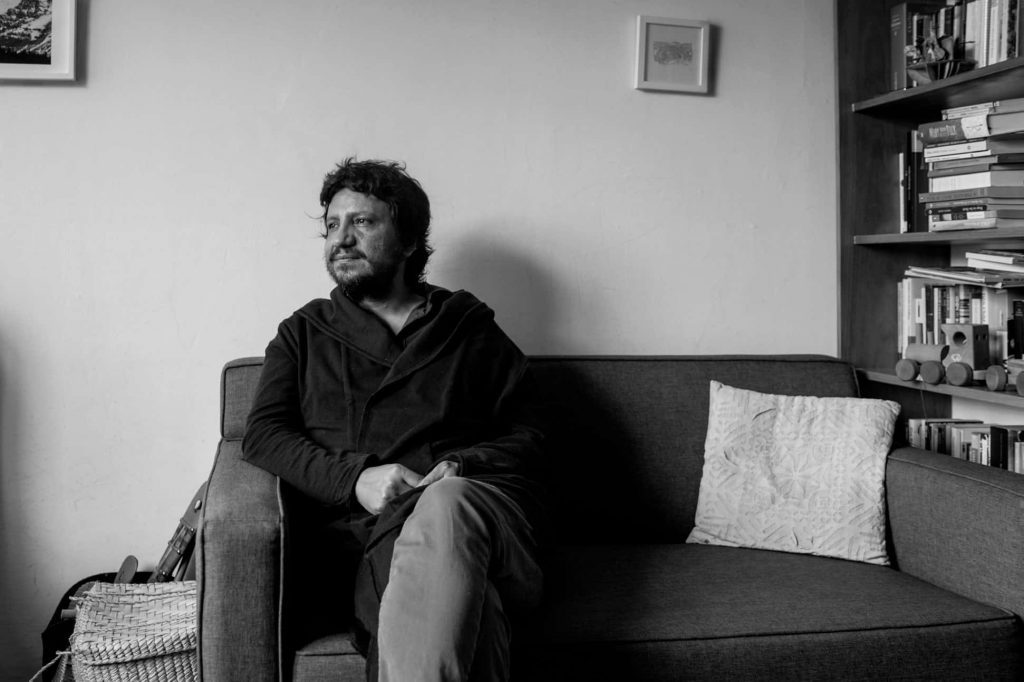 Alejandro Zambra sentado en un sofá gris, en blanco y negro, mirando hacia un lado en interior minimalista
