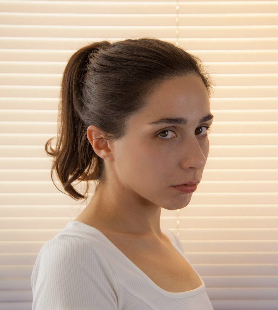 Retrato de perfil de mujer joven con cabello castaño recogido, vistiendo camiseta blanca sobre fondo de persiana beige