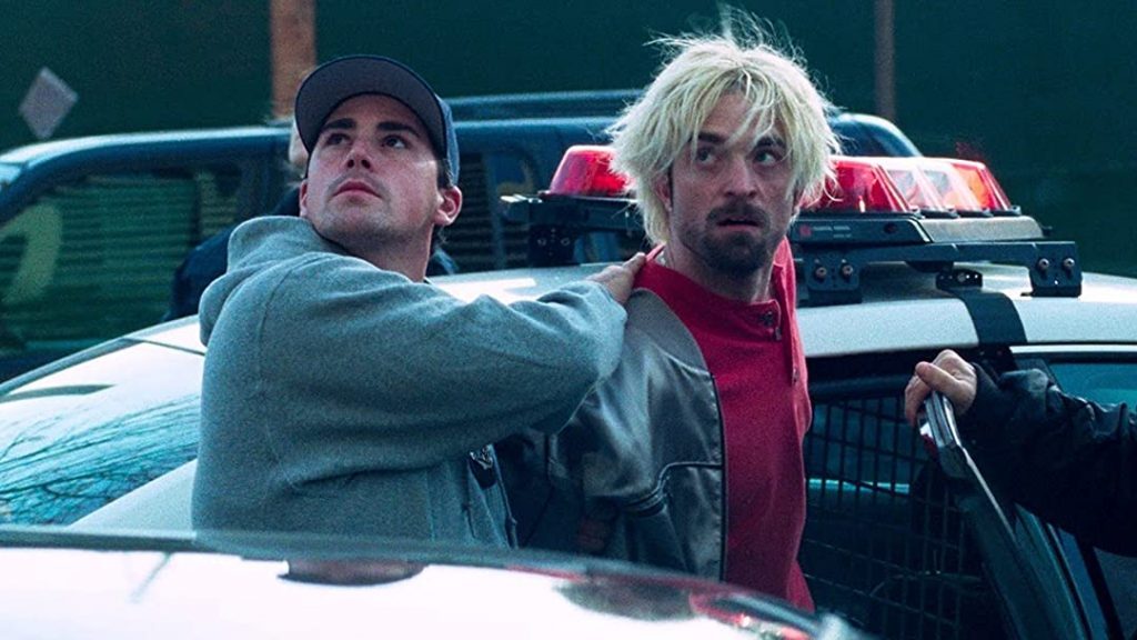 Dos hombres en un automóvil descapotable; uno de cabello rubio platinado viste en rosa, el otro en azul gris, ambos