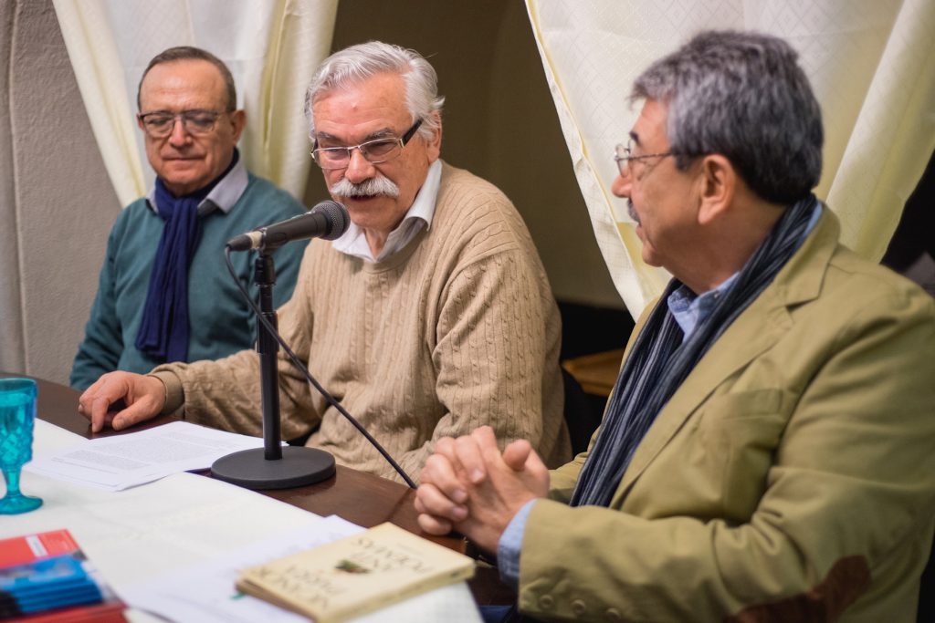 Sergio Infante y dos hombres en panel de conversación frente a micrófono durante evento cultural