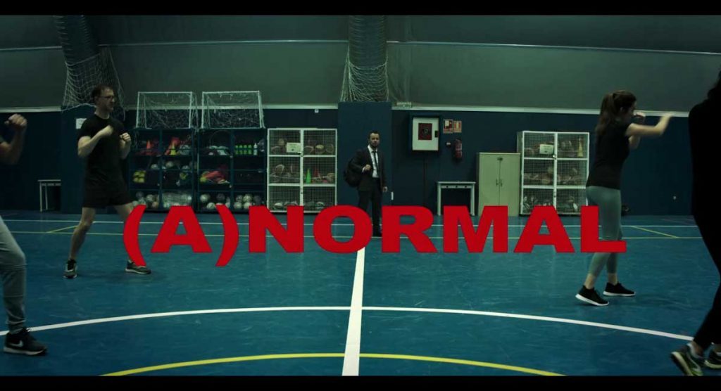 Escena de cancha de futsal con pared verde y texto rojo "ANORMAL" superpuesto, jugadores en movimiento durante partido