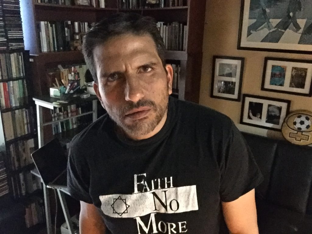 Cristián Brito Villalobos en su estudio, vistiendo camiseta negra con diseño blanco, rodeado de libros y fotografías