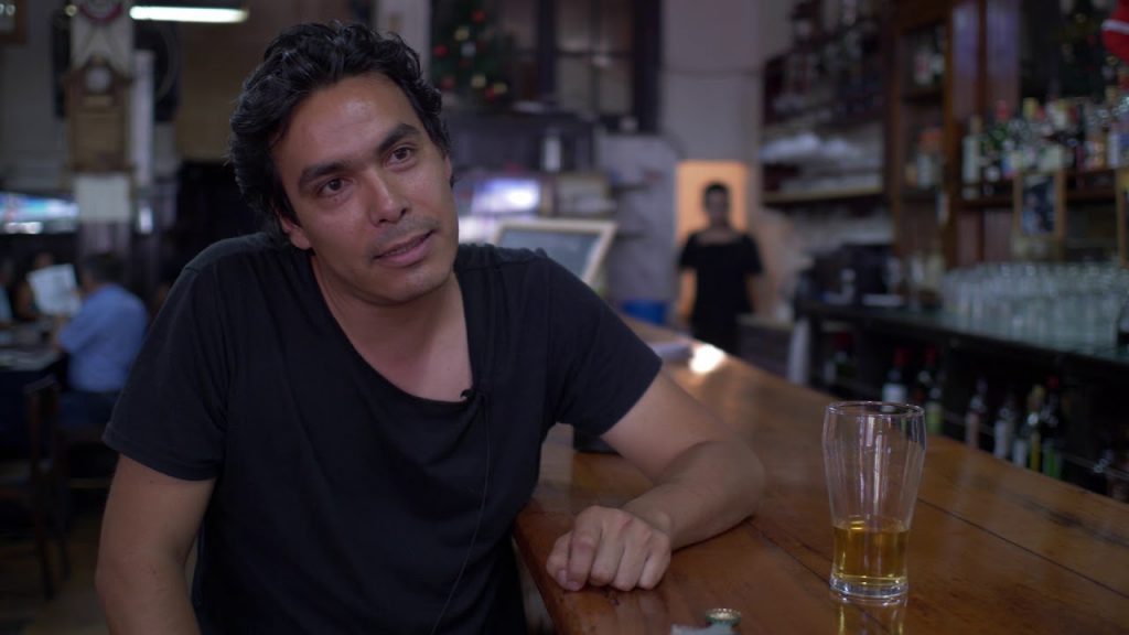 Cristián Geisse recostado en una barra de bar, vistiendo polera negra, con vaso de bebida clara frente a él en ambiente