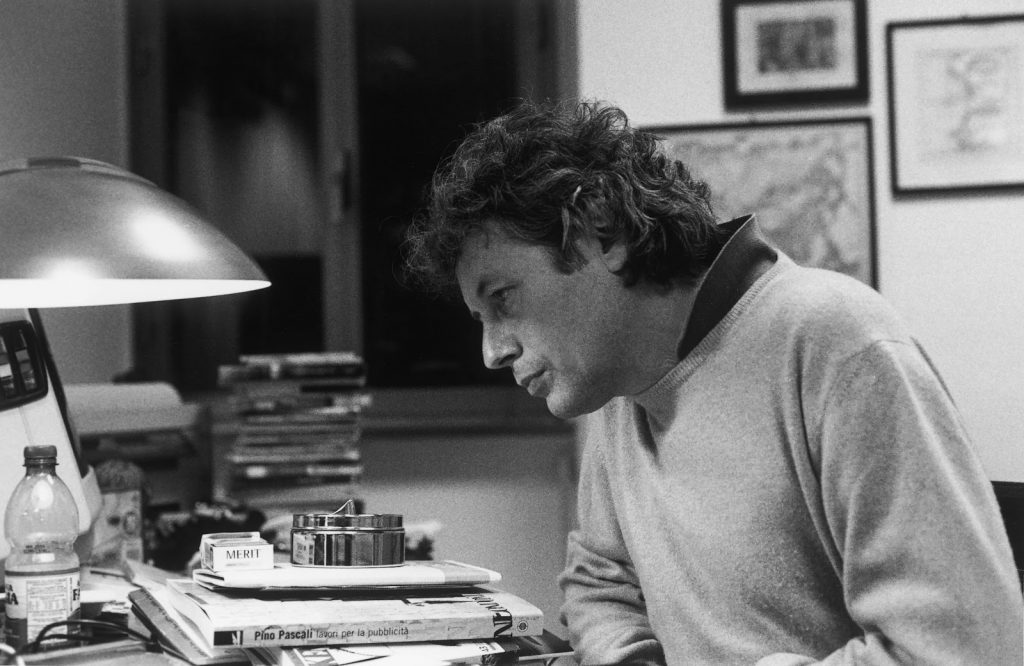 Alessandro Baricco sentado en su escritorio, rodeado de libros y papeles, bajo la luz de una lámpara de trabajo