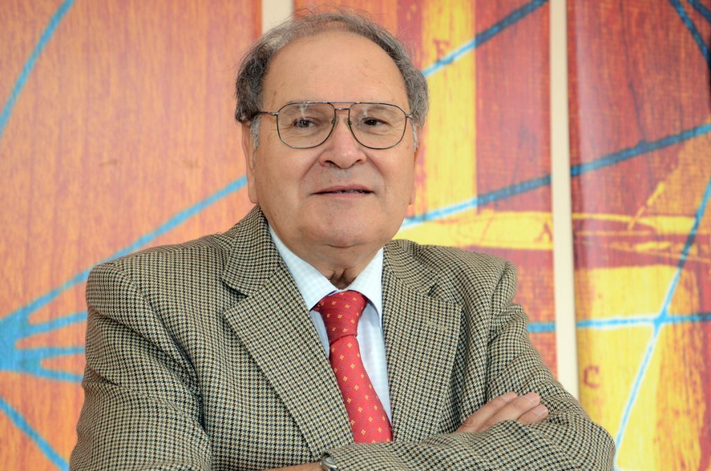 Jorge Pinto Rodríguez, historiador chileno con anteojos y traje gris, frente a fondo colorido