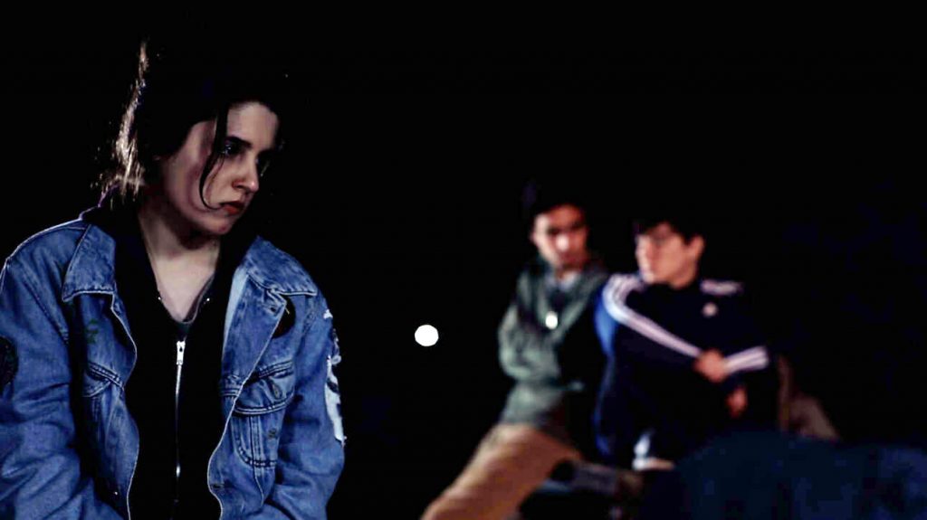 Hombre de lentes y chaqueta de mezclilla en primer plano; figuras borrosas al fondo en escena nocturna